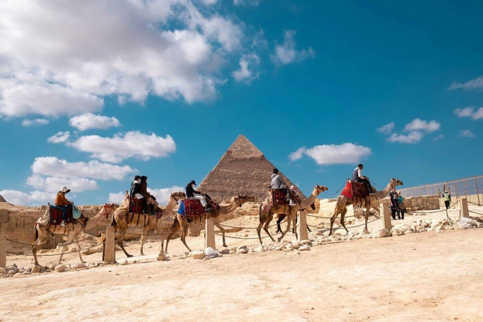 A WeRoad group trip riding a line of camels in the desert, with a large pyramid in the background under a blue sky.