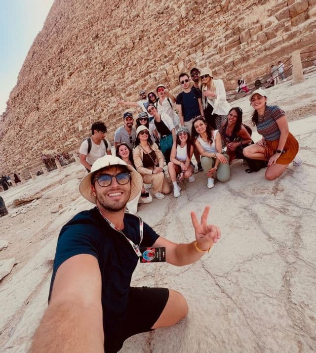 A man takes a selfie with a WeRoad group trip as they pose together on the large stone blocks at the base of a pyramid.