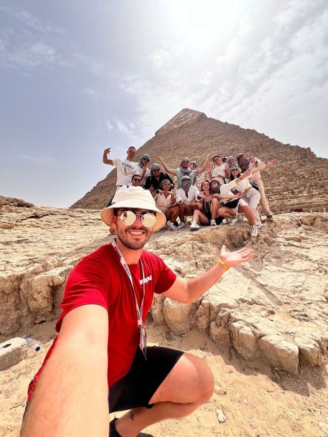 A man in a red shirt takes a selfie with a WeRoad group trip posing on sandy rocks in front of a large pyramid.