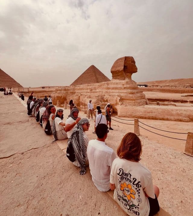 A WeRoad group trip sits in a line on a ledge, viewing the Great Sphinx with the pyramids of Giza in the background.