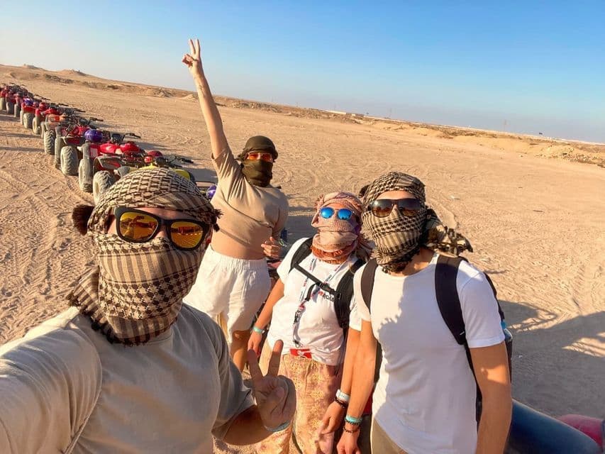 A WeRoad group trip wearing keffiyehs and sunglasses takes a selfie in the desert, with a line of quad bikes behind them.