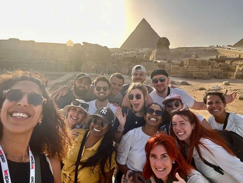 A WeRoad group trip takes a smiling selfie in front of the Great Pyramid and Sphinx under a golden sun.