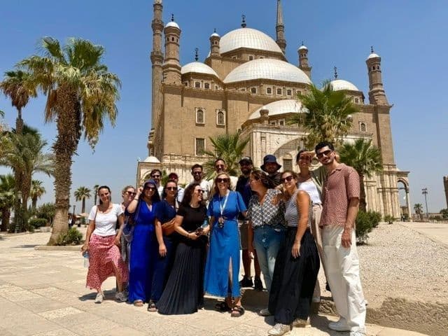 A WeRoad group trip posing for a photo in a sunny courtyard, with a large mosque with domes and minarets in the background.