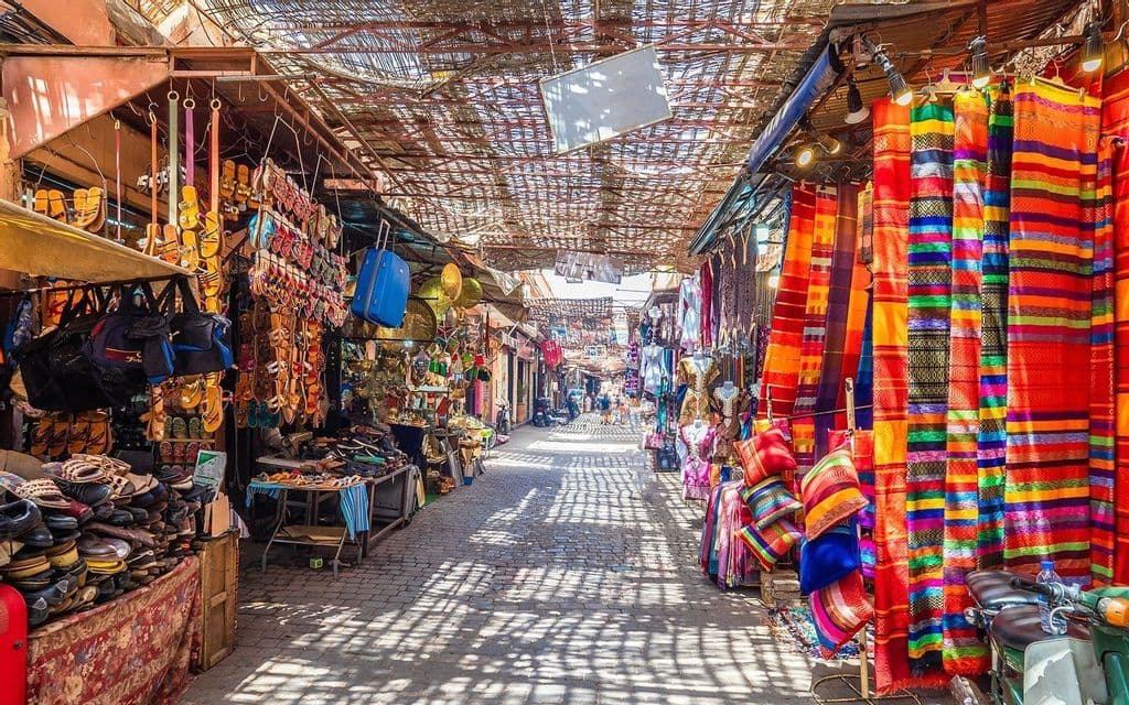A sunlit alley in a bustling bazaar, with market stalls on both sides displaying colorful textiles, bags, and handmade sandals.