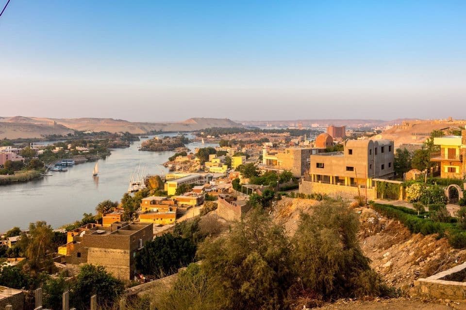 A high-angle view of a town built along a wide river, with sailboats on the water and a desert landscape in the background.
