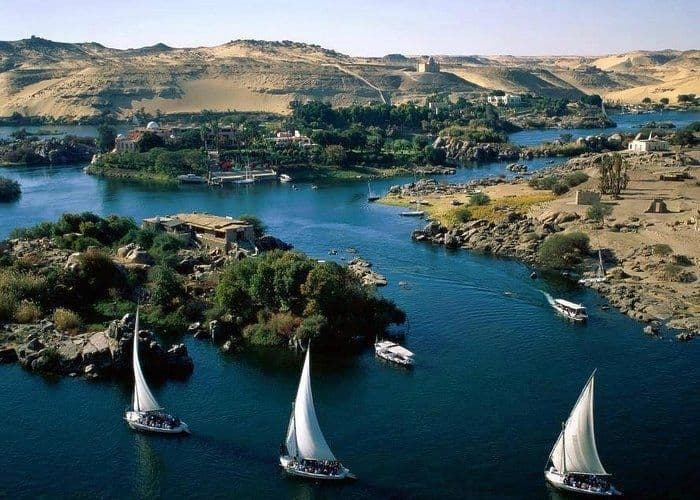 An aerial view of a WeRoad group trip on several white sailboats navigating a wide blue river between desert hills and green islands.
