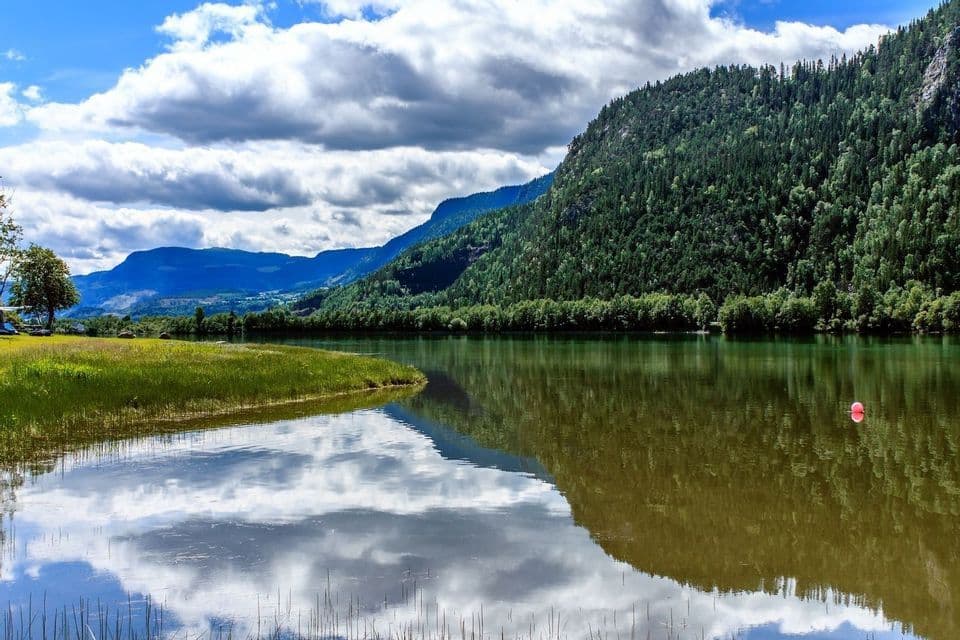 A calm lake reflects green, forested mountains under a cloudy blue sky.