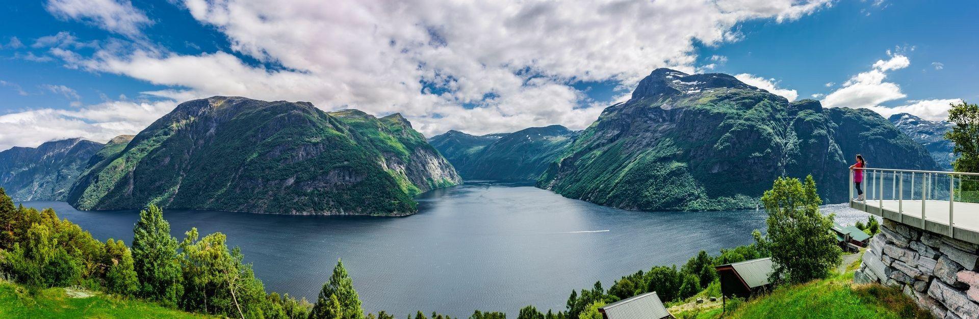 A person on an observation deck overlooks a vast fjord between steep green mountains under a partly cloudy sky.