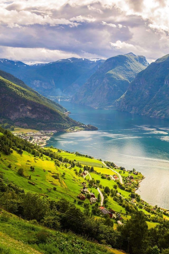 A high-angle view of a village on a green hillside along a fjord, with mountains and snow-capped peaks in the background.