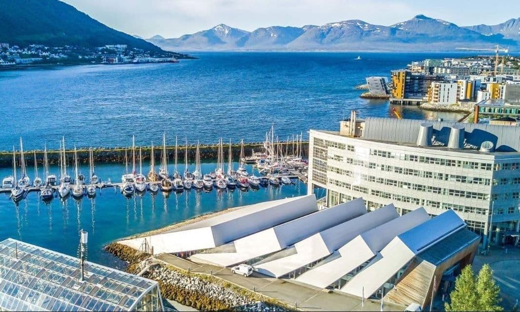 An aerial view of a modern harbor filled with sailboats, with a city and snow-capped mountains in the background.