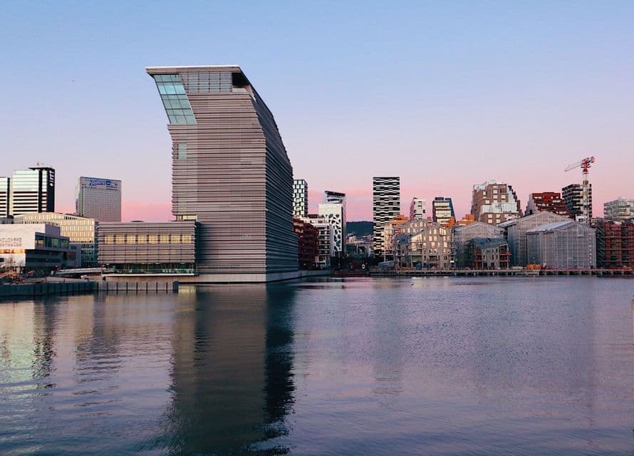 A modern city skyline with a distinctive leaning building is reflected on the waterfront at sunset.