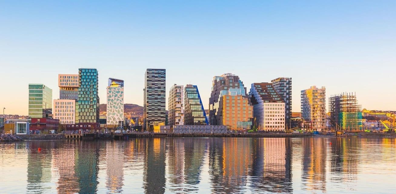 A modern city skyline with diverse architectural styles is reflected on the calm water of a waterfront at sunset.