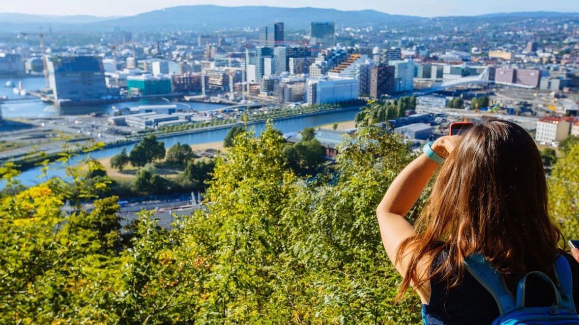 A woman with a backpack, seen from behind, photographs a sprawling city with a river from a high viewpoint.