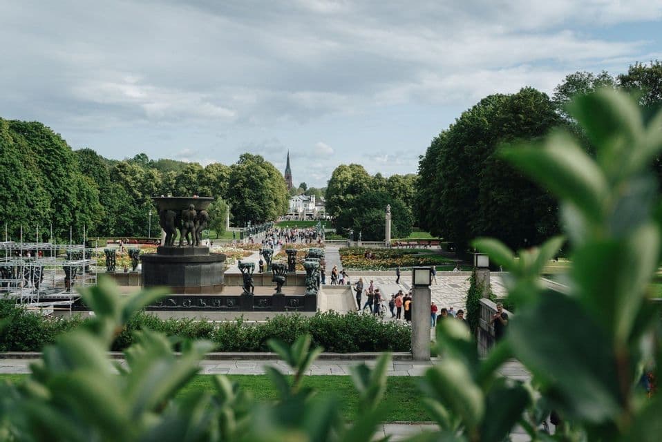 A bustling public park with people walking along a path lined with bronze statues and fountains, framed by green trees.