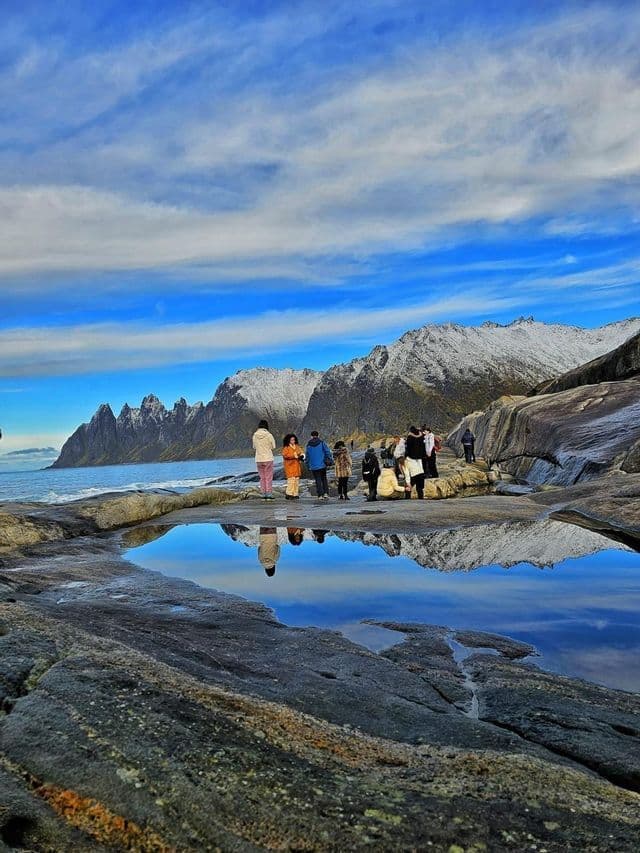A WeRoad group trip stands on a rocky shore, with snow-capped mountains and the sky reflected in a large tide pool.