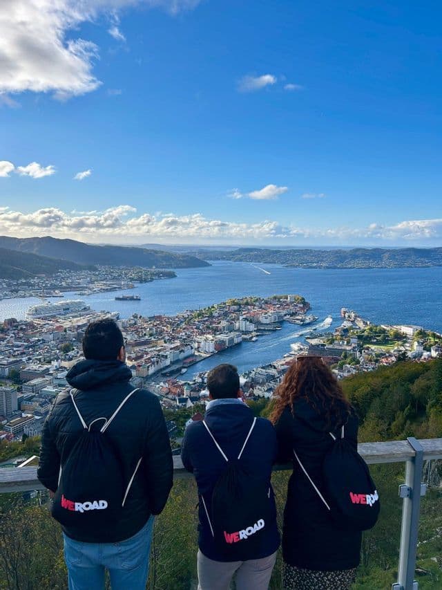 A WeRoad group trip of three people seen from behind, looking out from a viewpoint over a coastal city and fjord.
