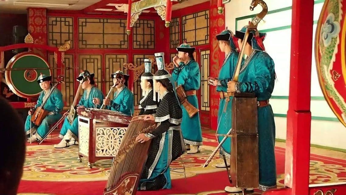 A group of musicians in traditional Mongolian attire play string and percussion instruments on an ornate indoor stage.