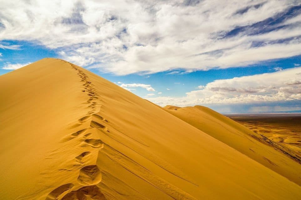 A trail of footprints leads up the ridge of a large, golden sand dune under a blue sky with white clouds.