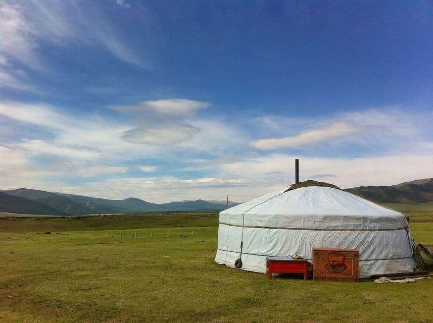 A white yurt stands in a green field with mountains in the distance under a blue sky with scattered clouds.