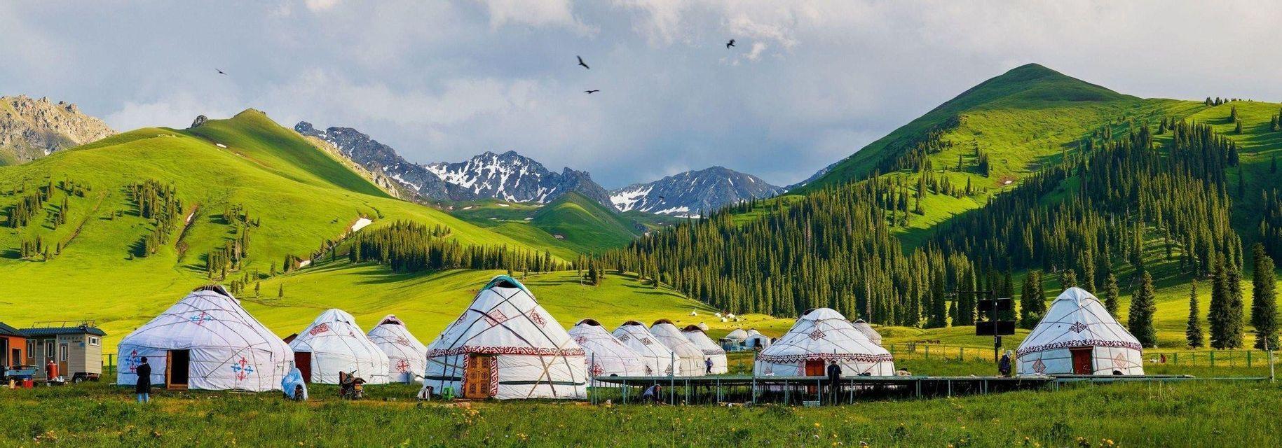 A village of traditional yurts in a grassy valley, with pine forests and snow-capped mountains in the background.