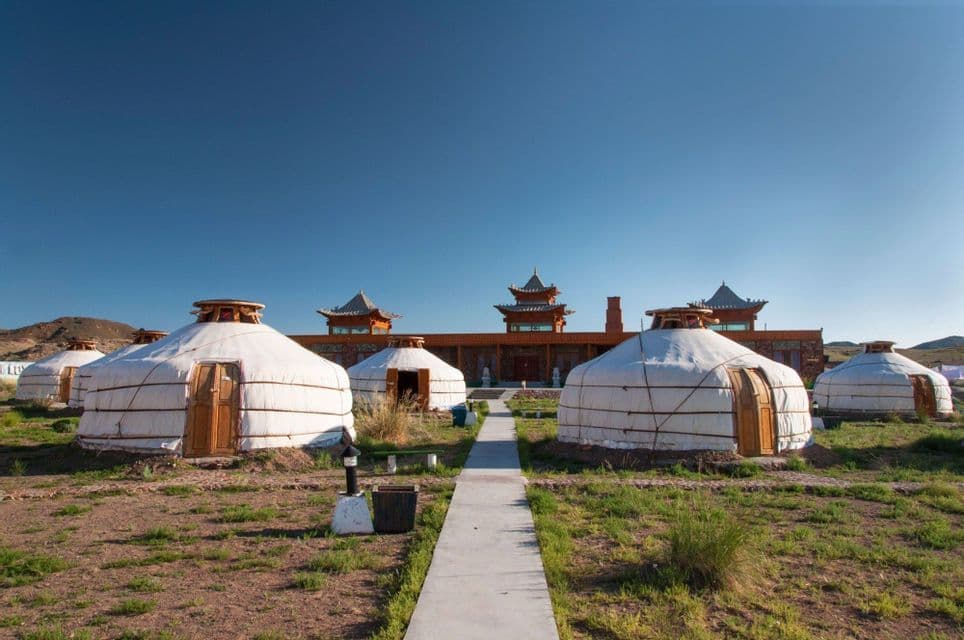 Several traditional white yurts stand in a grassy field in front of a large building with pagoda-style roofs under a clear sky.