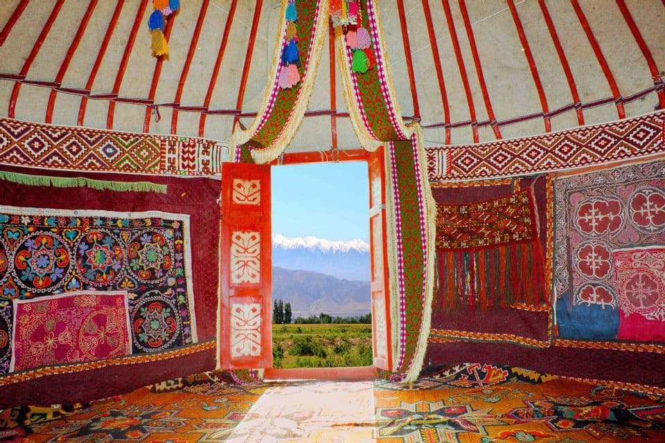 The interior of a colorful, traditional yurt with an open red door looking out onto a landscape of snow-capped mountains.
