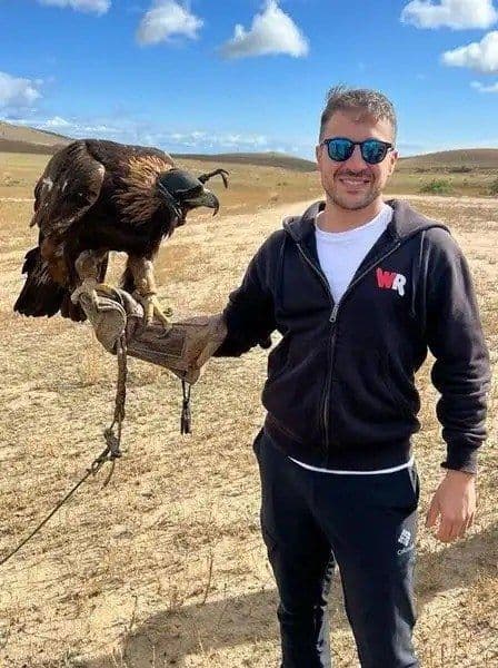 A smiling man wearing sunglasses holds a hooded golden eagle on his gloved hand in a dry, grassy field.