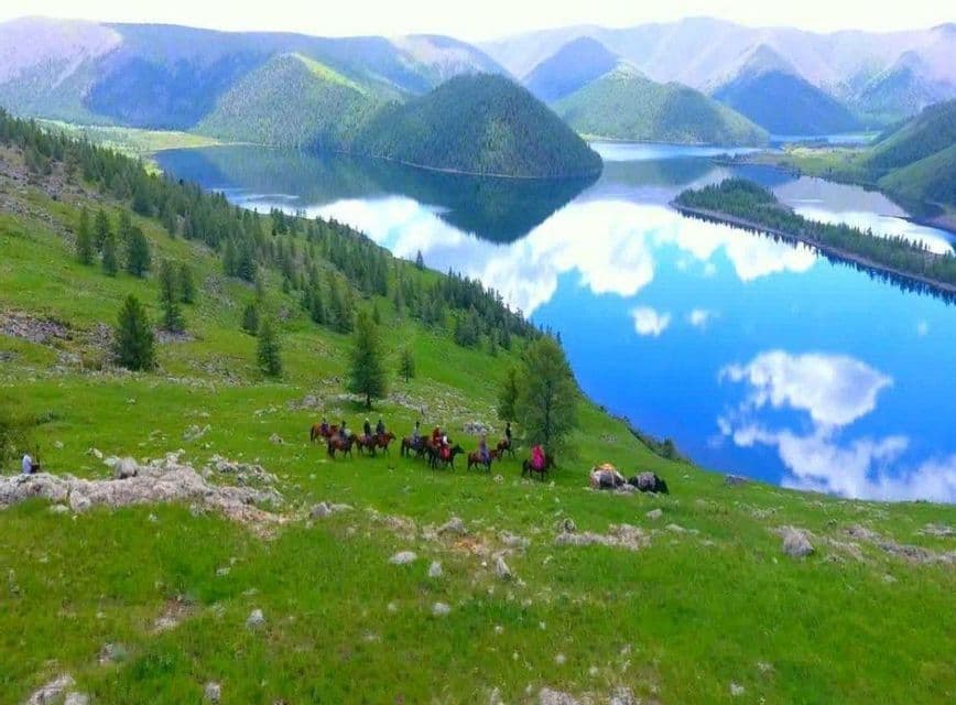 A WeRoad group trip on horseback travels along a green hillside overlooking a calm lake reflecting the cloudy sky.