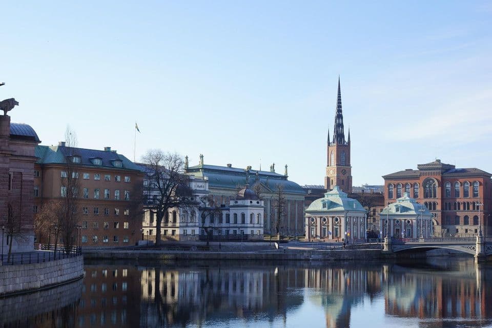 Uno scorcio urbano con edifici storici e un campanile di chiesa che si riflettono nell'acqua placida di un canale in una giornata di sole.