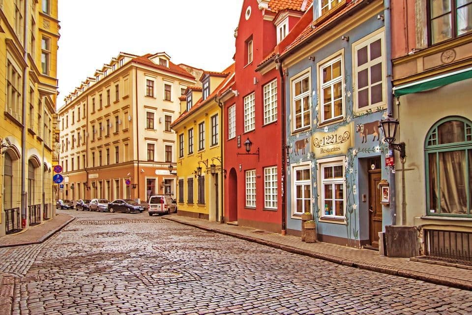 Una strada acciottolata serpeggiante, costeggiata da edifici colorati e storici e auto parcheggiate sotto un cielo coperto.