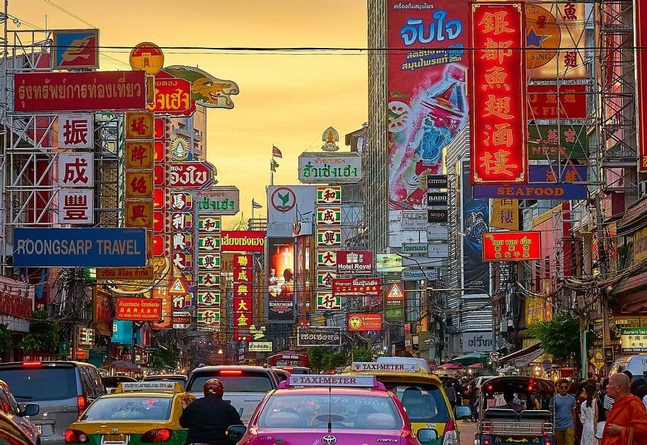 Una trafficata strada cittadina è piena di auto e taxi, affiancata da edifici ricoperti da innumerevoli insegne colorate sotto un cielo caldo e arancione.