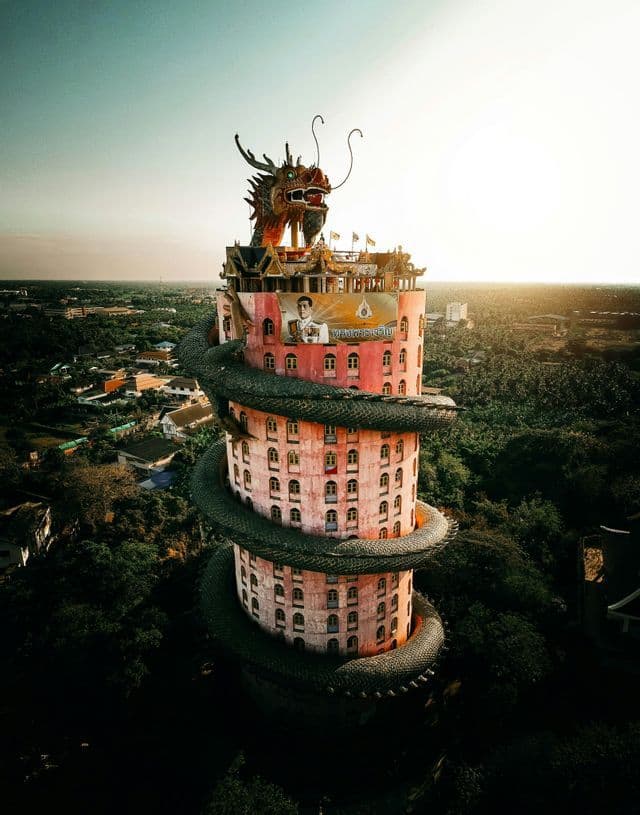 Una scultura di un drago gigante e scuro si avvolge attorno a un alto edificio cilindrico rosa, circondato da una foresta lussureggiante al tramonto.