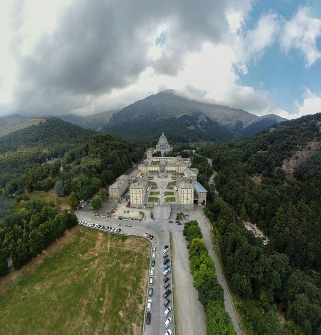 Veduta aerea di un vasto complesso di santuari con una cupola imponente, immerso in una valle montana boscosa sotto un cielo nuvoloso.