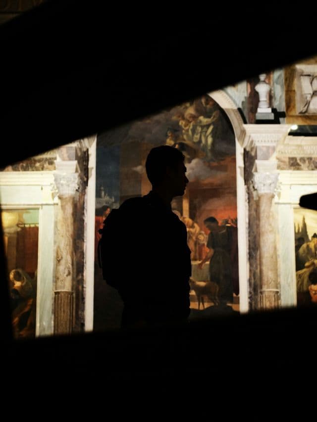 Una sagoma con zaino osserva un grande murale classico in un ambiente poco illuminato.