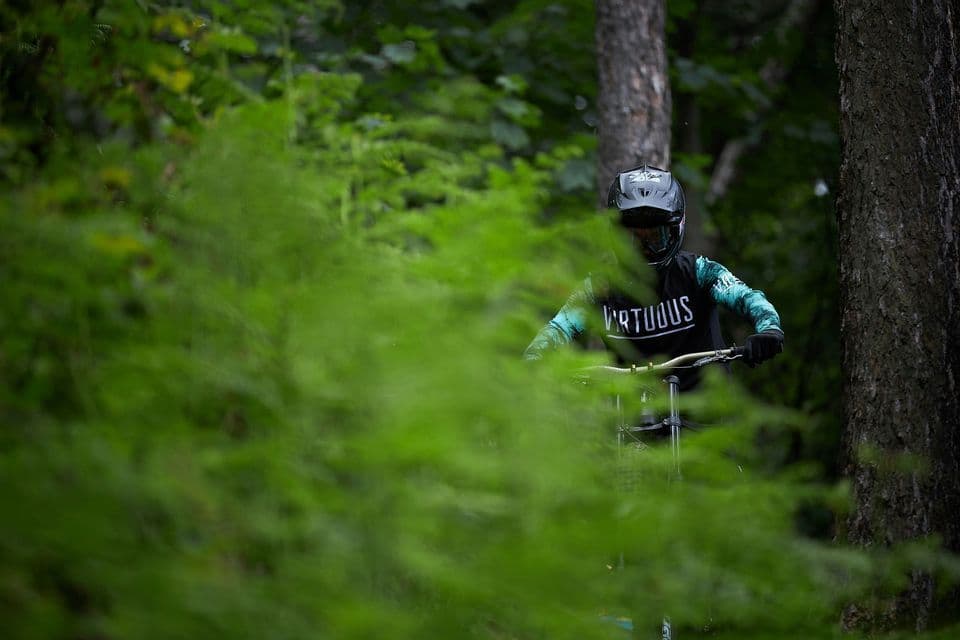 Un mountain biker con casco integrale e maglia attraversa una fitta foresta, visto da dietro il fogliame verde sfocato.