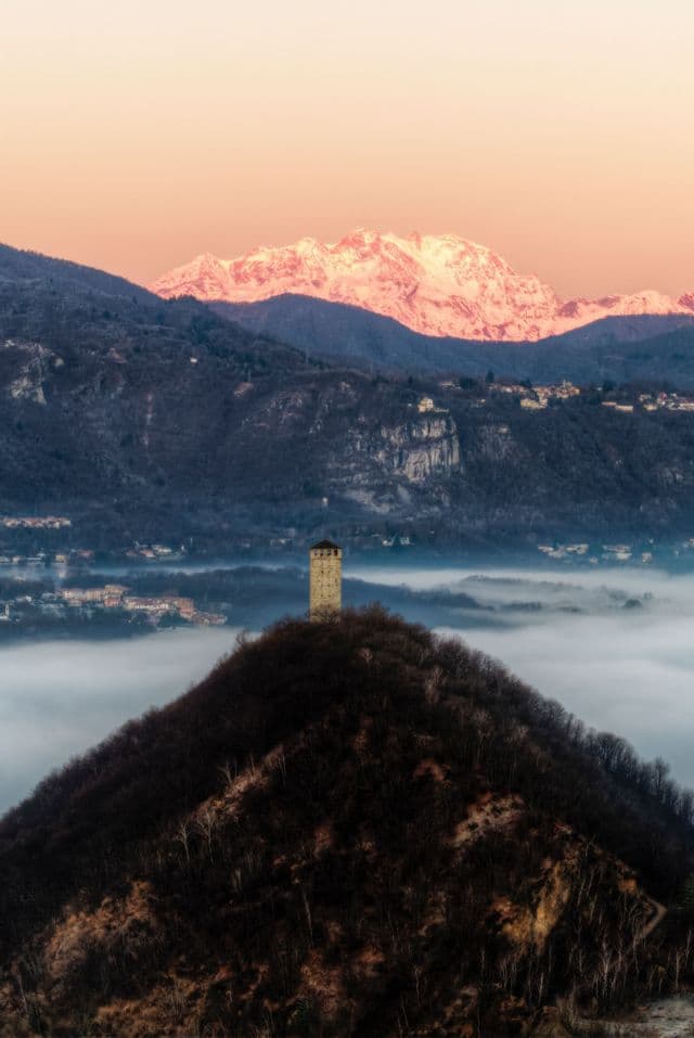 Una torre di pietra su una collina si erge sopra un mare di nebbia, con montagne innevate illuminate dal sole sullo sfondo.
