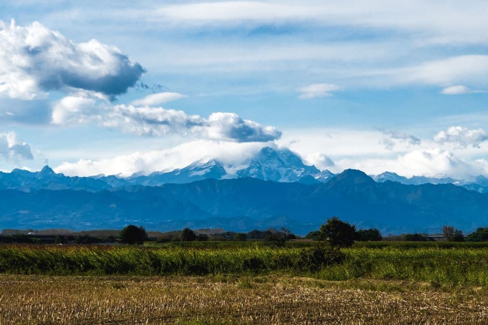 Una lontana catena montuosa innevata è visibile oltre campi verdi e gialli sotto un cielo blu nuvoloso.