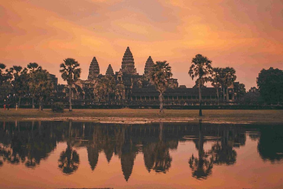 An ancient stone temple with spires and palm trees is reflected in a still body of water during a warm-hued sunset.