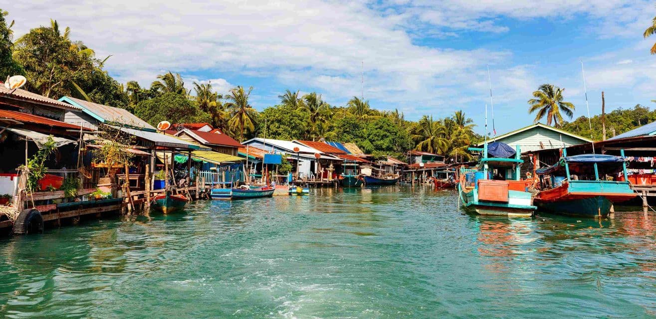 Coloridos barcos de pesca atracados junto a casas sobre pilotes en un pueblo ribereño con un exuberante bosque tropical de fondo.