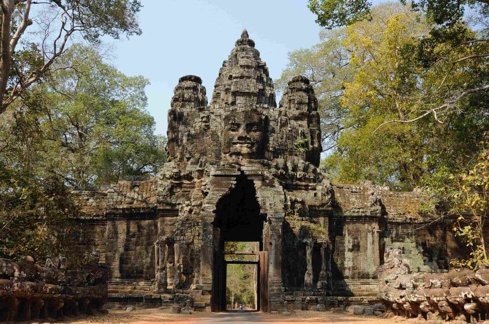 Ein antikes Steintor mit einem großen, gemeißelten Gesicht obenauf, das in einem Wald unter klarem Himmel steht.