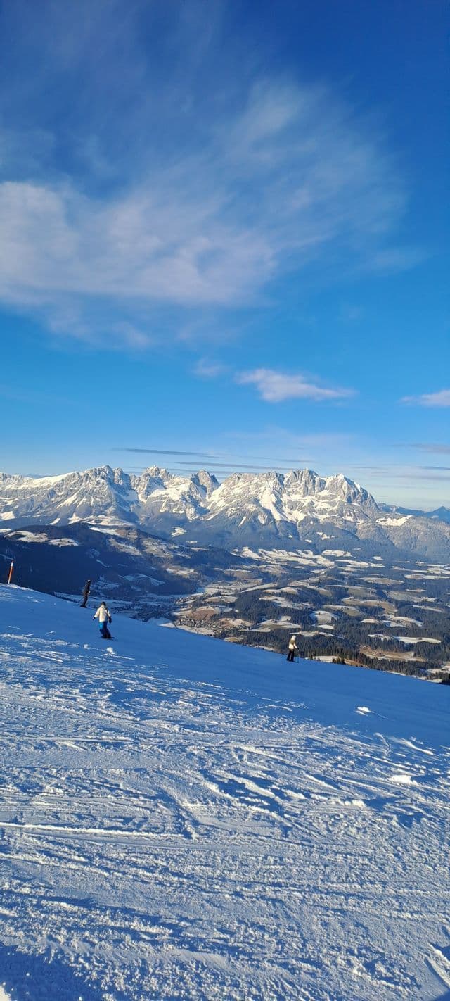 Skifahrer fahren einen schneebedeckten Hang hinab mit Blick auf ein weites Tal und schneebedeckte Gipfel unter einem blauen Himmel.