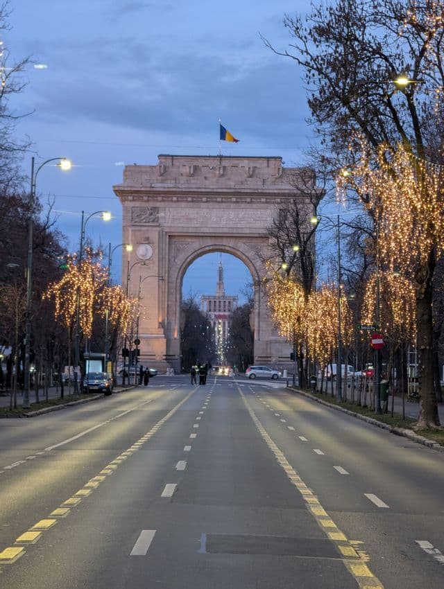 Ein Triumphbogen steht am Ende eines breiten Stadtboulevards, der in der Dämmerung von beleuchteten Bäumen gesäumt ist.