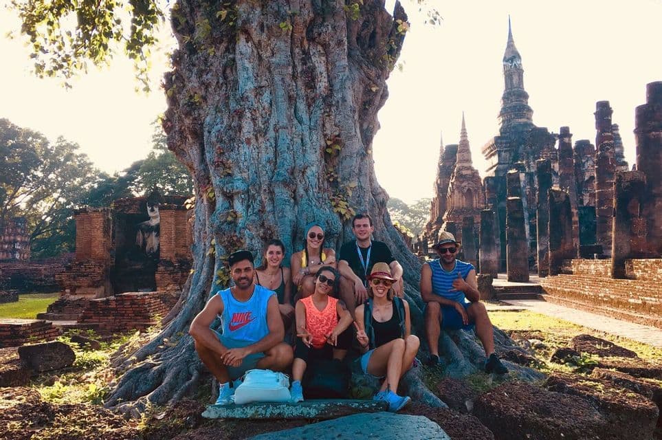 Un groupe WeRoad en voyage posant pour une photo au pied d'un grand arbre, avec des ruines de temples anciens et une statue de Bouddha en arrière-plan.