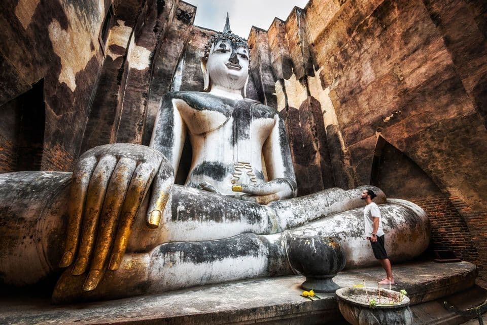 A man stands looking up at a giant, weathered stone Buddha statue enclosed by towering brick walls.
