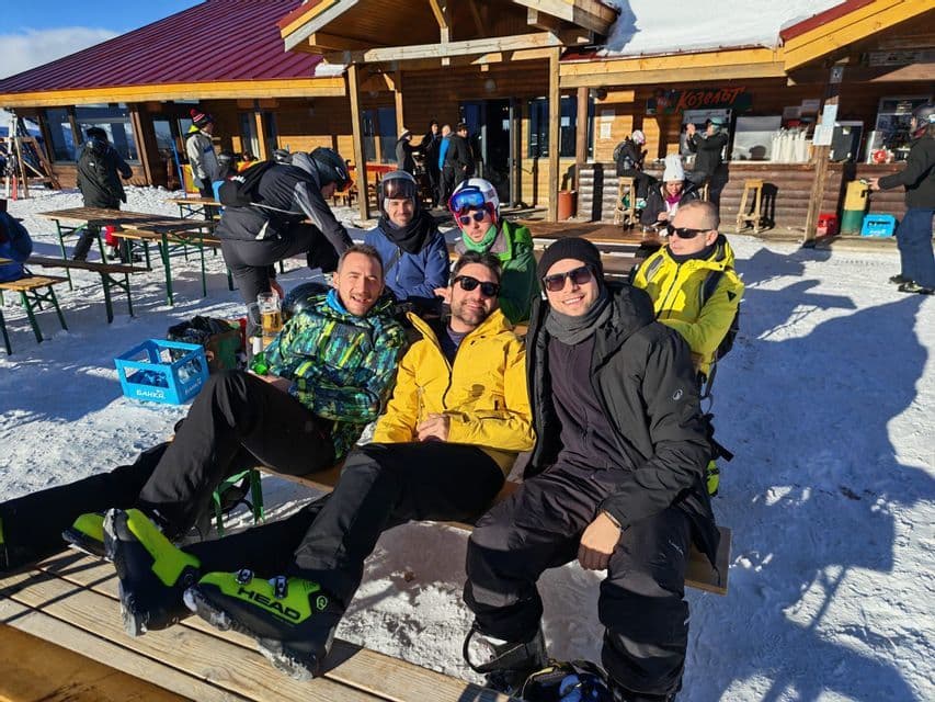 Eine WeRoad-Gruppe von Leuten entspannt in Skikleidung auf Holzbänken vor einer Berghütte im Schnee.