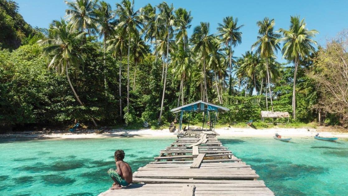 A person sits on a weathered wooden pier extending over turquoise water towards a beach lined with palm trees.