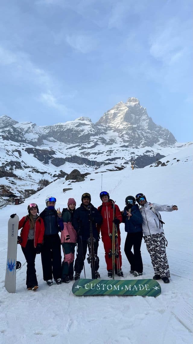 Eine WeRoad-Gruppe posiert mit Skiern und Snowboards auf einem schneebedeckten Hang vor einem felsigen Berggipfel.
