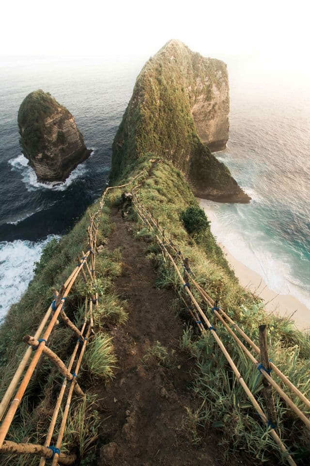 A narrow dirt path with a bamboo fence runs along a steep, grassy ridge overlooking a turquoise ocean and a white sand beach.