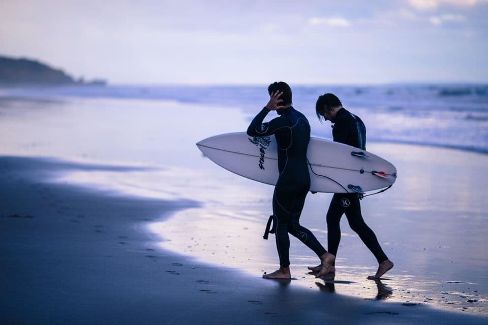 Two surfers in wetsuits walk on a wet beach at dusk, carrying a surfboard along the shoreline.