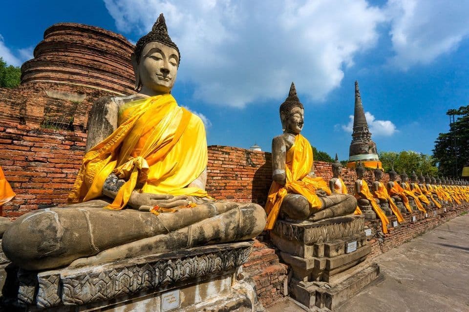 Eine Reihe alter steinerner Buddha-Statuen, in gelbe Safrangewänder gehüllt, sitzt vor Ziegelstupas unter blauem Himmel.